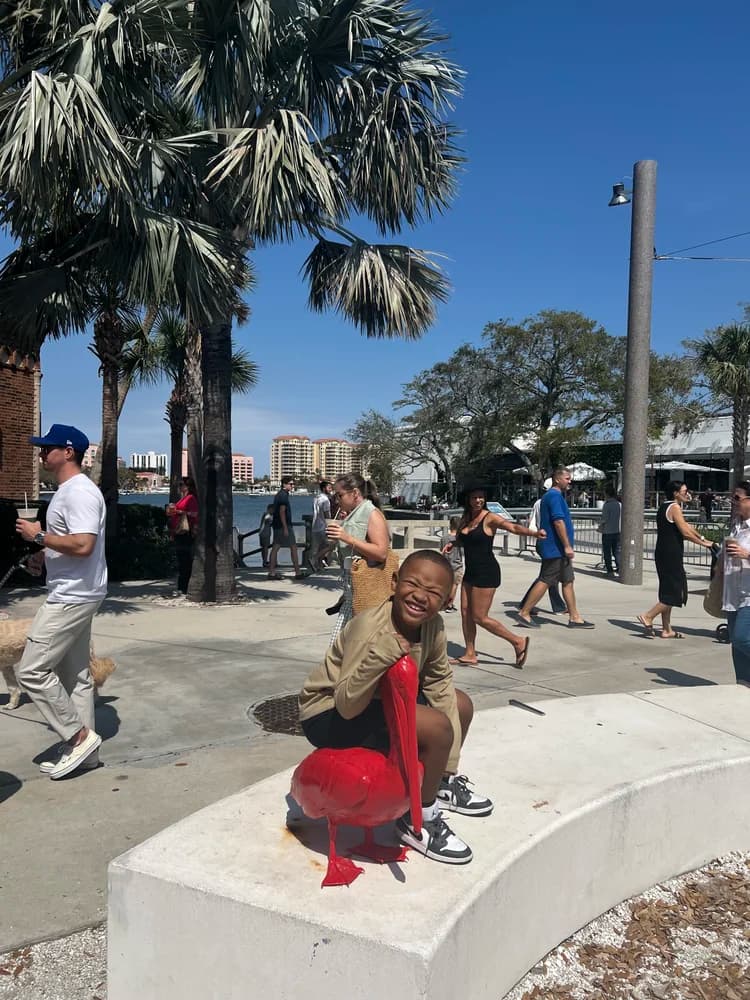 Junior sitting on a red pelican statue along a waterfront promenade